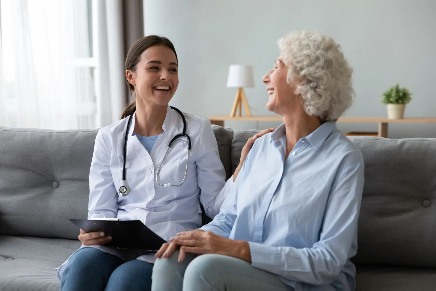 Friendly young nurse in white coat holding clipboard laughing with elderly female patient during homecare visit consultation Friendly young nurse in white coat holding clipboard laughing with elderly female patient during homecare visit consultation