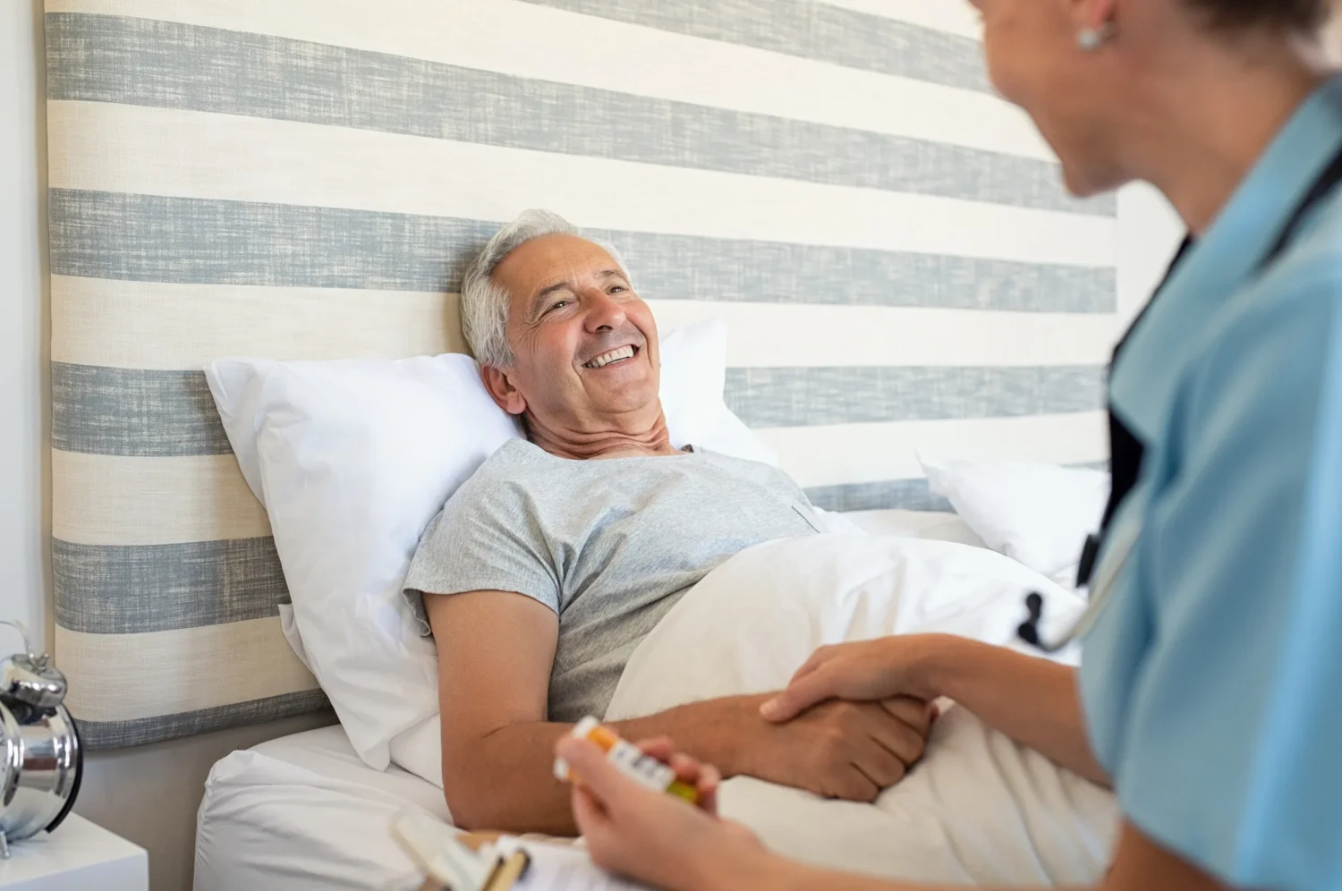 Nurse holds the hand of a patient lying in bed, discussing medication, in a bright, simple hospital room with a striped wall Nurse holds the hand of a patient lying in bed, discussing medication, in a bright, simple hospital room with a striped wall